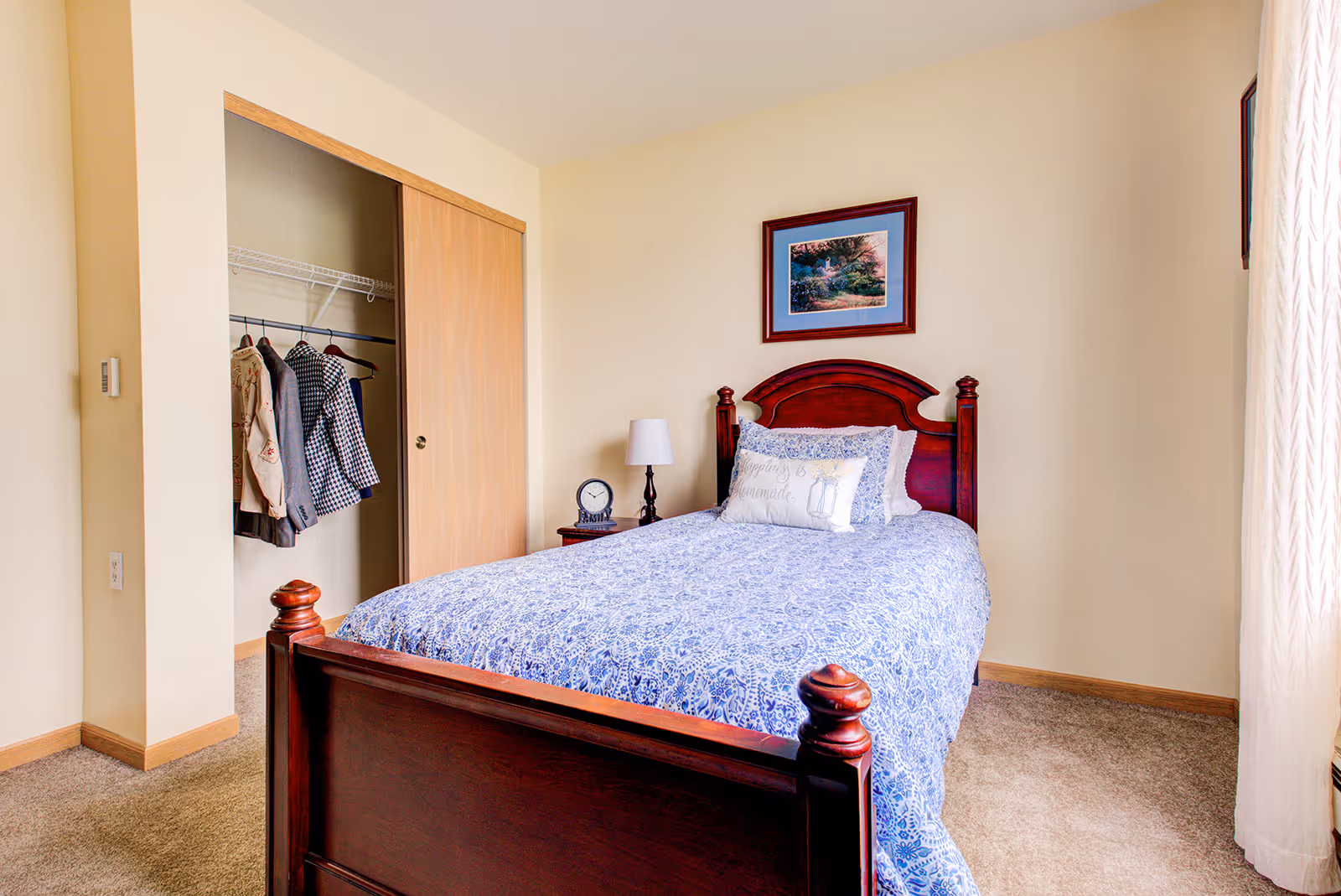 A bedroom with a wooden bed frame and a blue patterned bedspread. There is a pillow with the text 'Happiness is homemade' on the bed. Next to the bed is a small wooden nightstand with a lamp and a clock. A framed picture hangs on the wall above the bed. To the left, there is an open closet with sliding wooden doors and several jackets hanging inside. Light beige walls and carpeted floor complete the room.