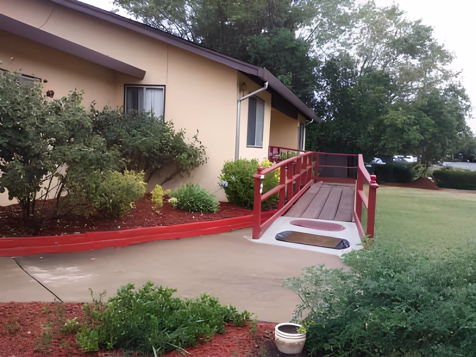 Exterior view of a beige building with a red wooden ramp leading to an entrance. The ramp has handrails on both sides and is surrounded by green bushes and trees. There is a concrete pathway and well-maintained landscaping with mulch and plants.
