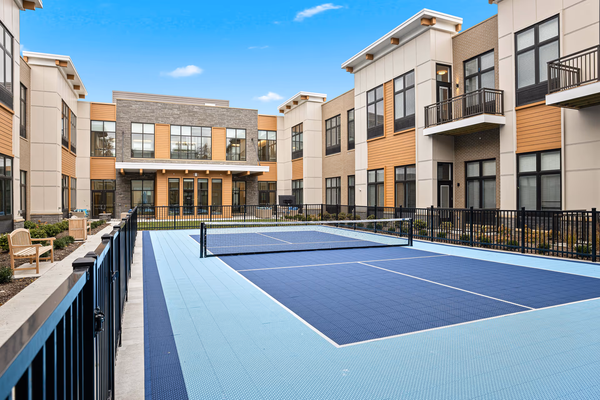 Outdoor courtyard area of The Ashton at Mayfield Heights featuring a fenced pickleball court with a net, surrounded by a two-story building with large windows and balconies under a clear blue sky.
