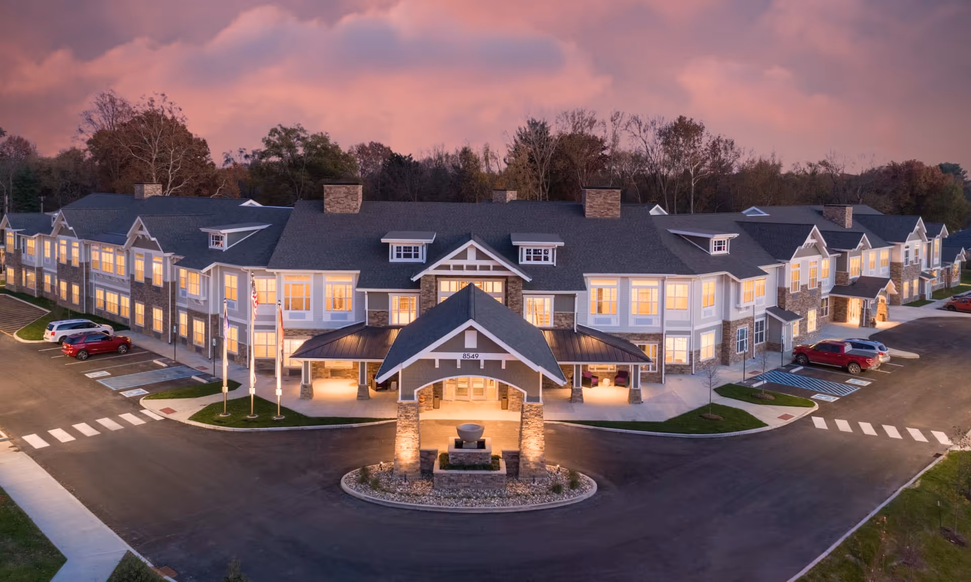 Exterior view of The Commons on Meridian senior living facility at dusk, showing a large two-story building with many lit windows, a covered entrance with stone pillars, a circular driveway, parked cars, and trees in the background under a pink and purple sky.