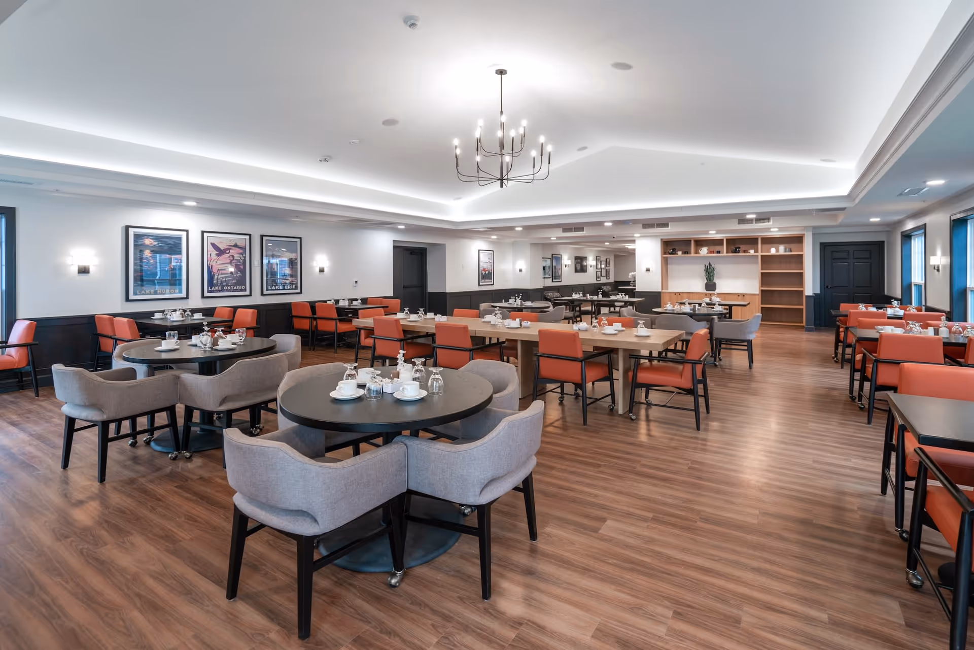 Spacious dining room with multiple tables and chairs arranged neatly. The room features a modern chandelier, wood flooring, framed artwork on the walls, and a combination of gray and orange upholstered chairs. Tables are set with cups, glasses, and napkins, ready for dining.