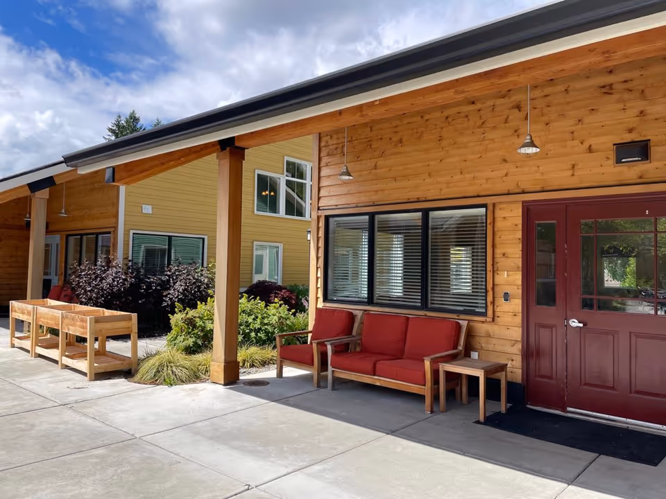 Outdoor covered patio area with wooden walls and a red door. There is a wooden bench with red cushions and a small wooden side table. Raised garden beds and various plants are visible along the walkway. The sky is partly cloudy.