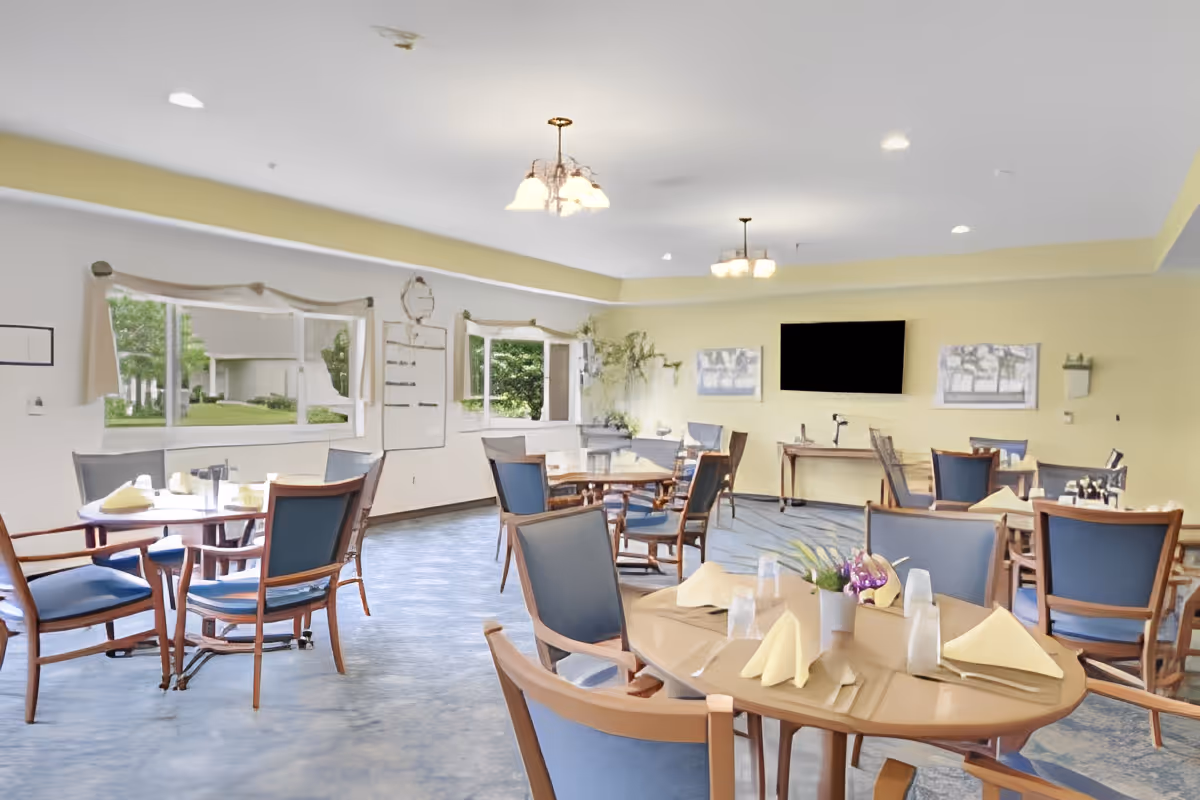 A bright dining room with multiple round tables set with folded yellow napkins, glasses, and a small flower arrangement. The room has large windows with views of greenery outside, blue cushioned wooden chairs, and a wall-mounted flat screen TV. The walls are painted light yellow and white, and the floor has a blue patterned carpet.
