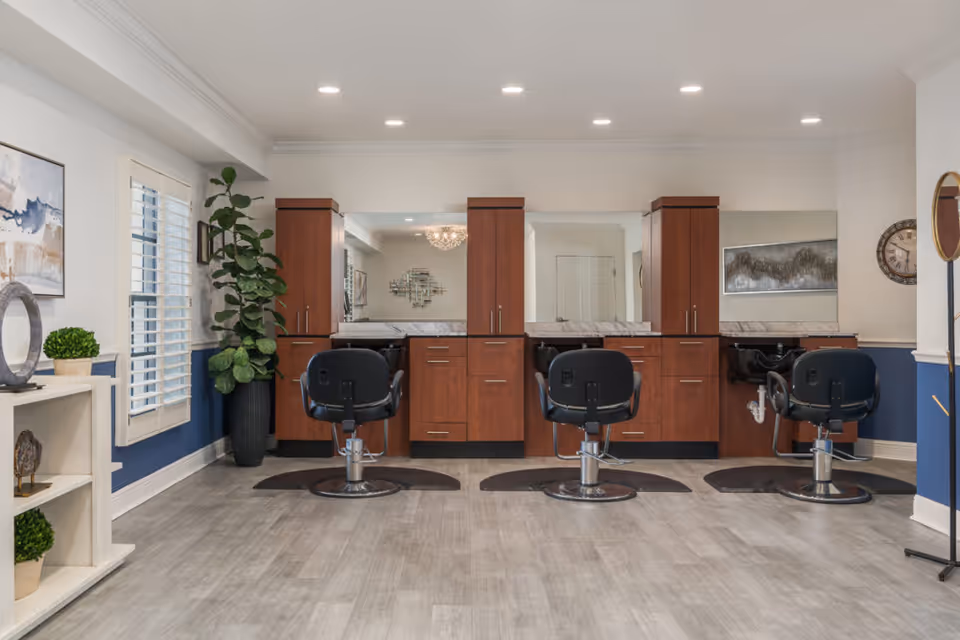Interior view of a hair salon area in a senior living facility with three black salon chairs in front of wooden cabinets and mirrors. The room has light gray flooring, white walls with blue wainscoting, a large potted plant, decorative artwork, and a clock on the wall.