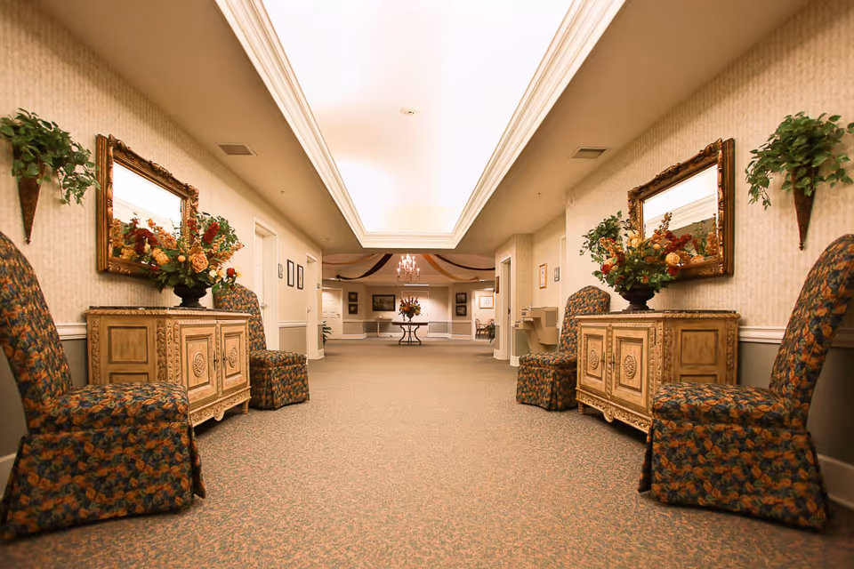 A well-lit hallway in a senior living facility with floral-patterned chairs and wooden cabinets topped with flower arrangements on both sides. Large mirrors hang above the cabinets, and the hallway leads to a room decorated with a chandelier and additional floral arrangements.