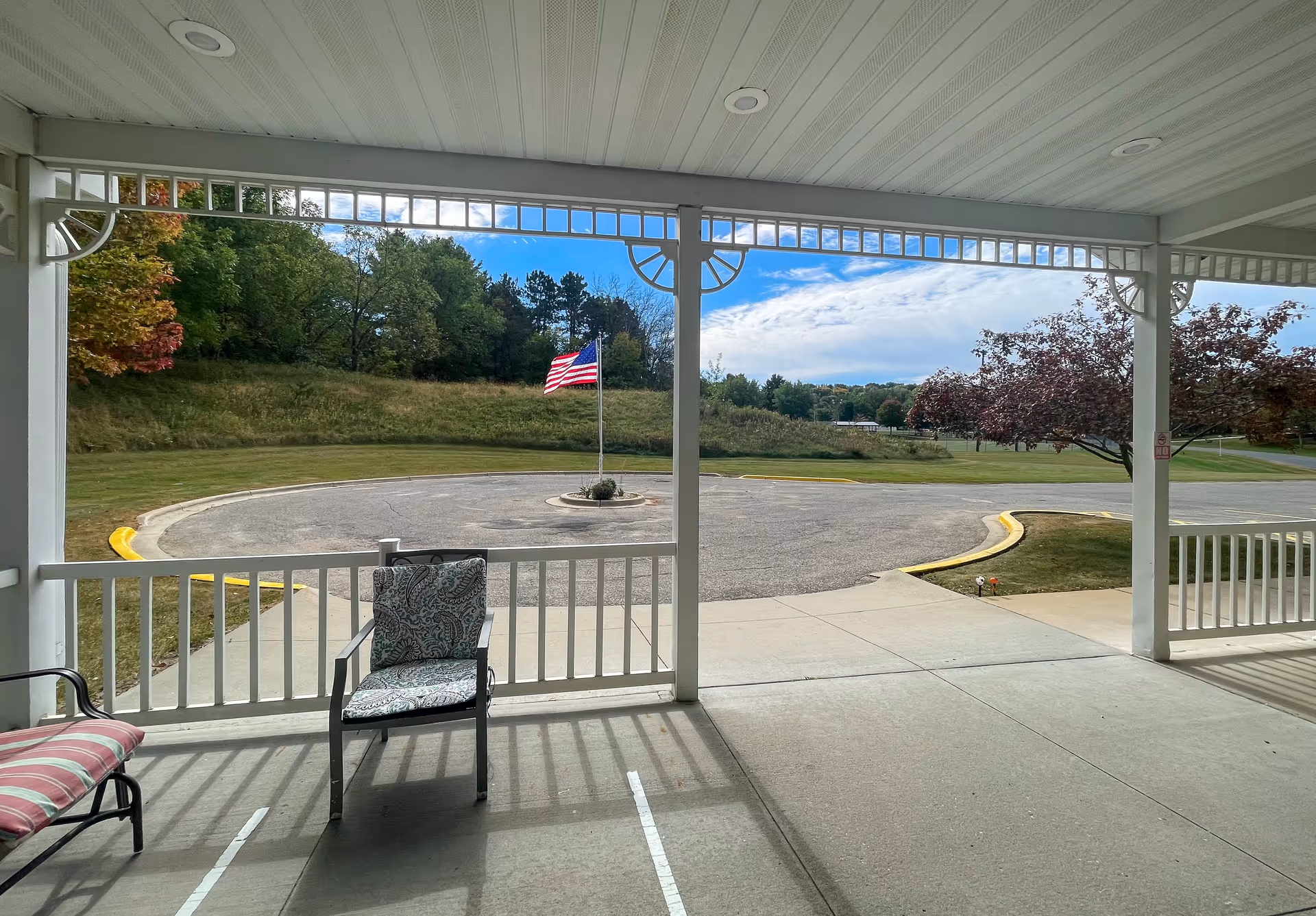 View from a covered porch area with white railings and ceiling, showing two cushioned chairs. Beyond the porch is a circular driveway with an American flag in the center, surrounded by grassy areas and trees under a partly cloudy sky.