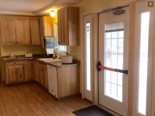 Interior view of a kitchen area with wooden cabinets, a countertop with a sink, a dishwasher, a toaster, and a crockpot. There is a glass door with a push bar exit leading outside, and windows on either side of the door allowing natural light in.
