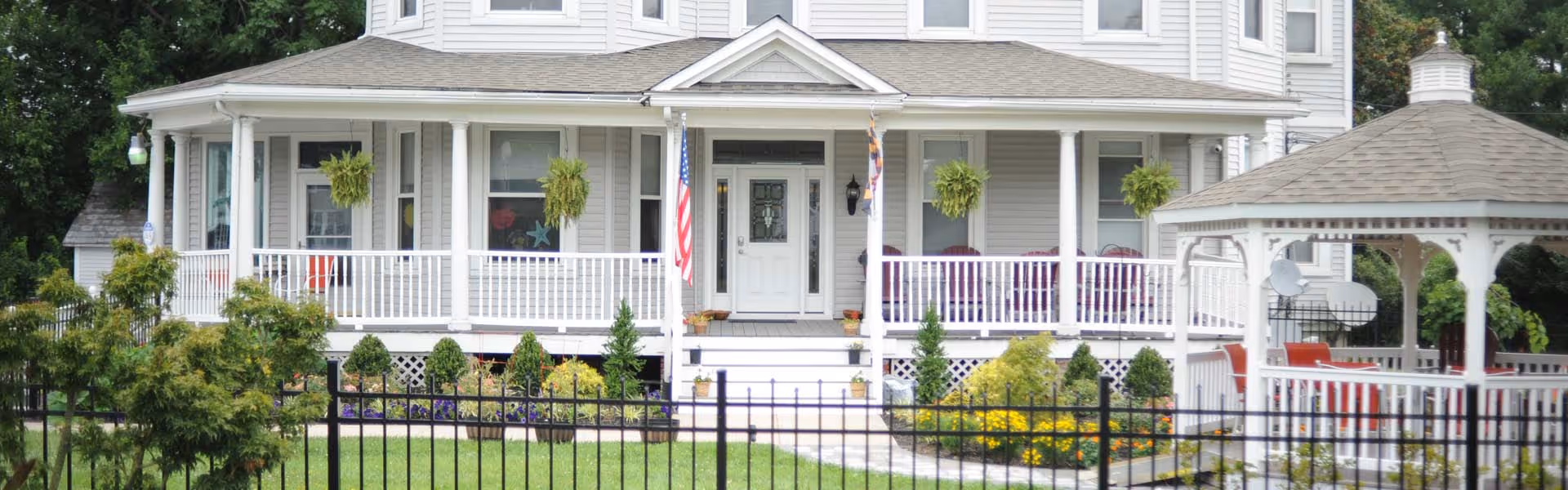 Front exterior view of a large, light gray, two-story house with a wraparound porch, white railings, hanging plants, and an American flag. The house is surrounded by a well-maintained garden with bushes, flowers, and a black metal fence. There is a white gazebo with red chairs on the right side of the house.