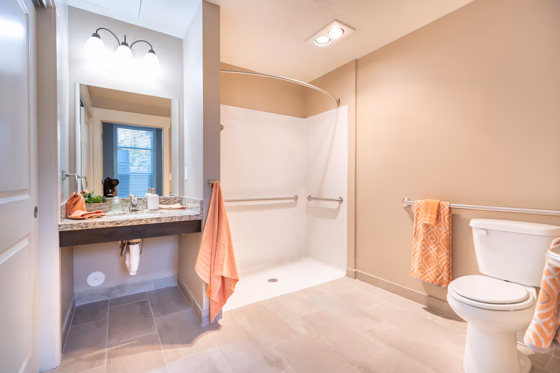 A clean and spacious bathroom with beige walls and light-colored tile flooring. The bathroom features a walk-in shower with grab bars, a white toilet with an orange patterned towel hanging on a grab bar, and a vanity with a granite countertop, a sink, and a mirror above it. There are orange towels hanging on the wall and countertop. The lighting is bright with three bulbs above the mirror and recessed ceiling lights.