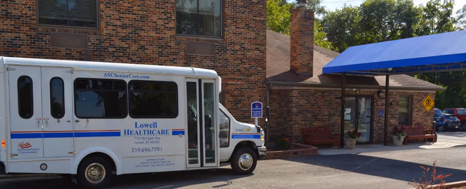 A white shuttle bus parked in front of a brick building with a blue awning at the entrance. The bus has Lowell Healthcare branding and contact information on its side. There are benches and potted plants near the building entrance, and several cars are parked in the background.