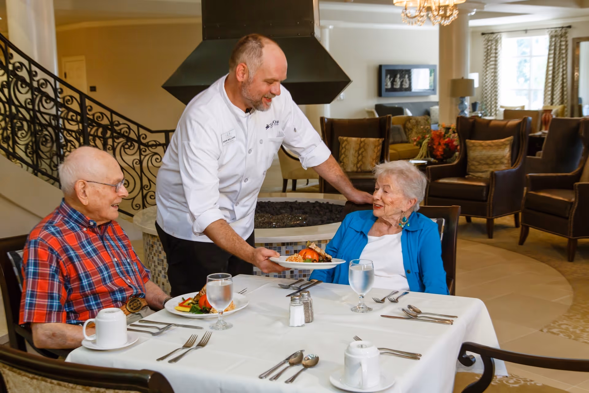 A chef serves a plated meal to two elderly residents seated at a table in a senior living dining/lounge area.