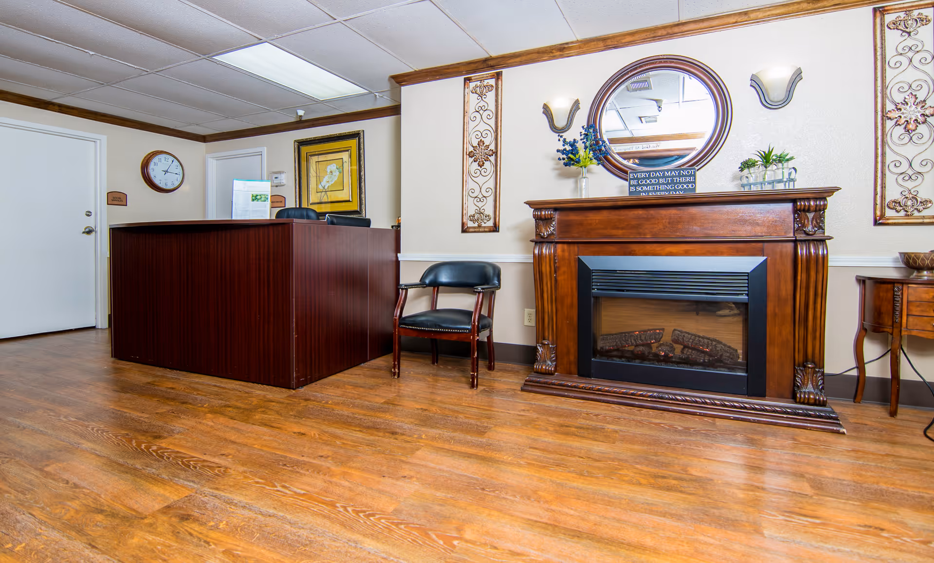 Interior view of a reception area in Sugar Land Health Care Center featuring a wooden reception desk, a black chair, a decorative wooden electric fireplace with a round mirror above it, wall sconces, and framed artwork on the walls. The floor is wooden and the ceiling has fluorescent lighting.