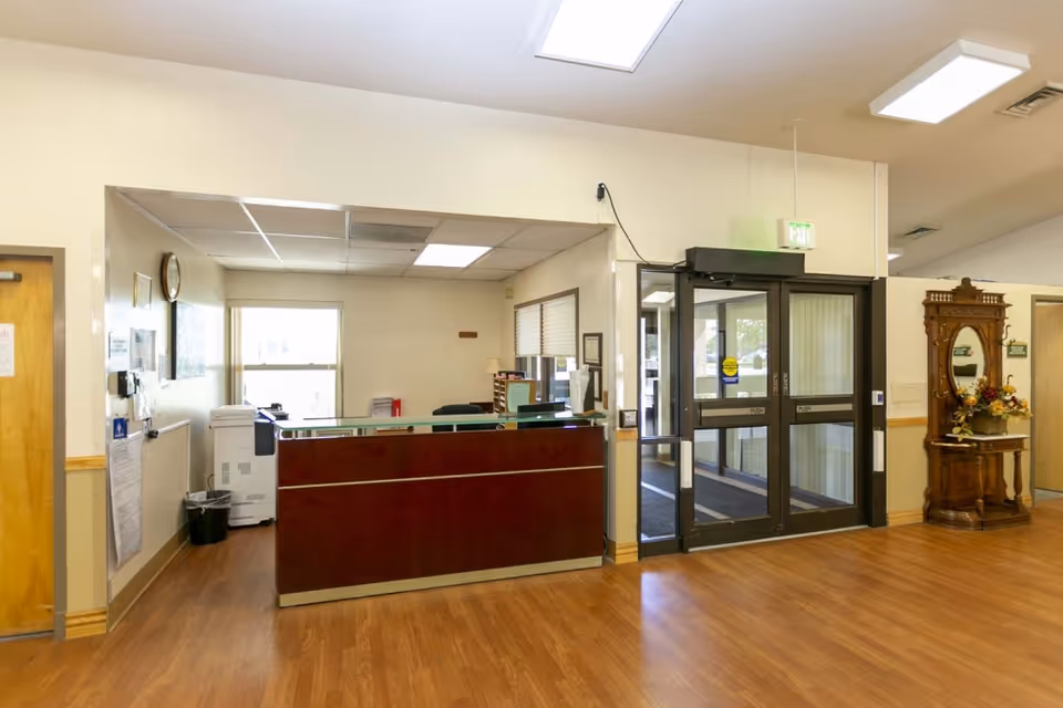 Reception area inside The Terrace at Mt. Ogden facility featuring a wooden front desk, glass entrance doors, a wooden floor, and a decorative wooden table with a mirror and flower arrangement on the right side.