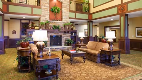 A spacious senior living facility common area with a high ceiling and balcony railing above. The room features a large stone fireplace decorated with plants, two beige sofas, wooden side tables with ornate lamps, and a coffee table with books. The carpet has a patterned design, and the walls are painted in warm tones with wood paneling accents.