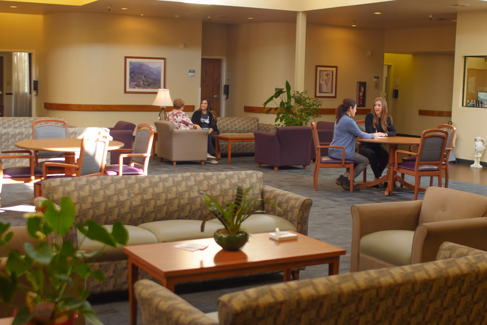 A spacious and well-lit common area with multiple seating arrangements including sofas, armchairs, and round tables with chairs. Four people are seated in pairs, engaged in conversation. The room has beige walls, framed artwork, a potted plant, and a table lamp, creating a warm and welcoming atmosphere.