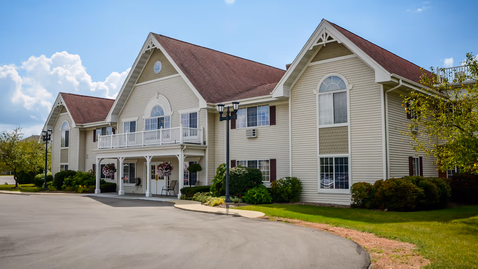 Exterior view of a two-story senior living facility building with beige siding and a red roof under a partly cloudy blue sky. The building features multiple large windows, a balcony, and a covered entrance with hanging flower baskets. There are bushes and trees around the building and a paved driveway in front.