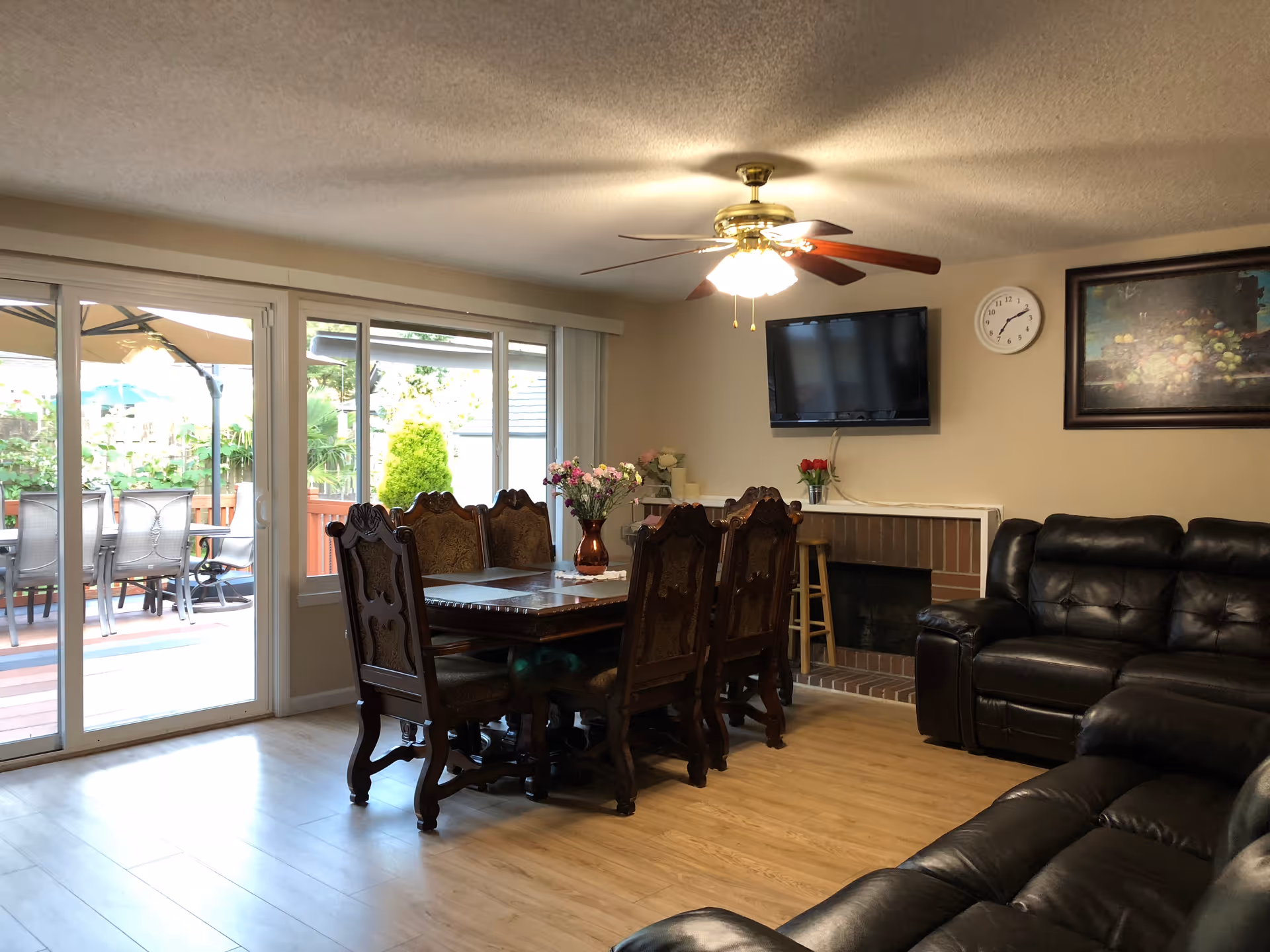 A cozy living and dining area with a wooden dining table and six ornate chairs in the center. There is a ceiling fan with lights above the table. To the right, there are black leather sofas arranged near a brick fireplace with a mounted flat-screen TV above it. A clock and a framed painting hang on the wall. Large sliding glass doors on the left open to an outdoor patio with a table and chairs under an umbrella. The room has light-colored wooden flooring and beige walls.