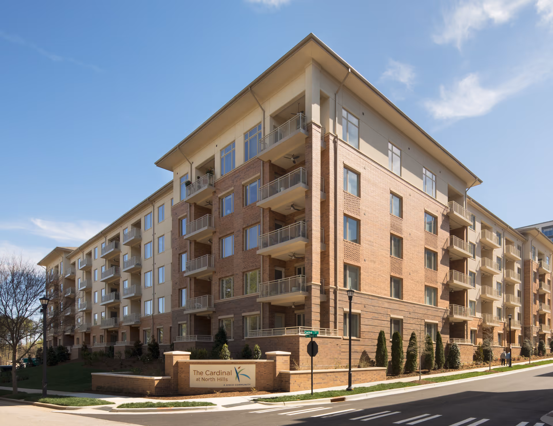Exterior view of a multi-story residential building with balconies, clear blue sky, and a sign in front that reads 'The Cardinal at North Hills'. The building has a mix of brick and light-colored siding with landscaped greenery around the base.