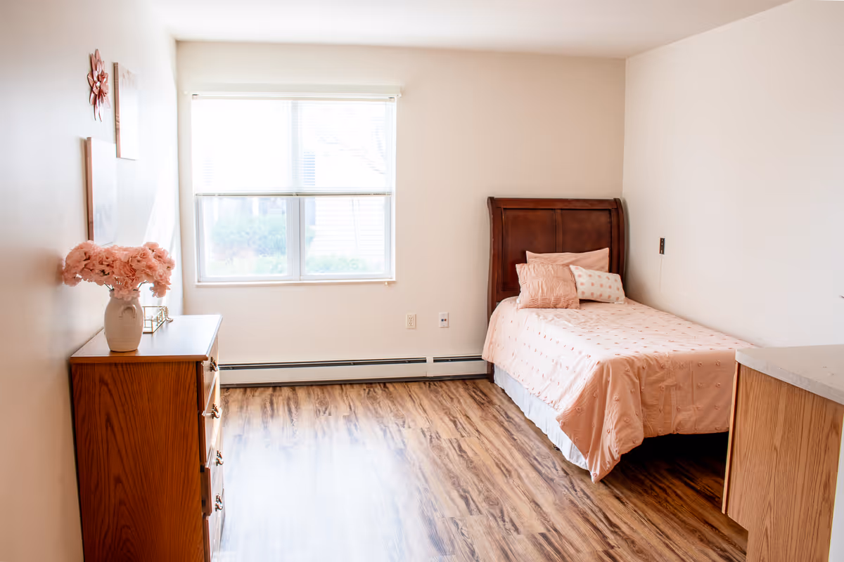 A bright and clean senior living bedroom with a single wooden bed adorned with pink bedding and pillows. There is a wooden dresser with a vase of pink flowers on top, and a large window letting in natural light. The room has light-colored walls and wood-patterned flooring.
