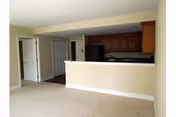 View of an empty living area with beige carpet and walls, adjacent to a kitchen with dark wood cabinets, black appliances, and a breakfast bar. There are two doors visible on the left side of the image.
