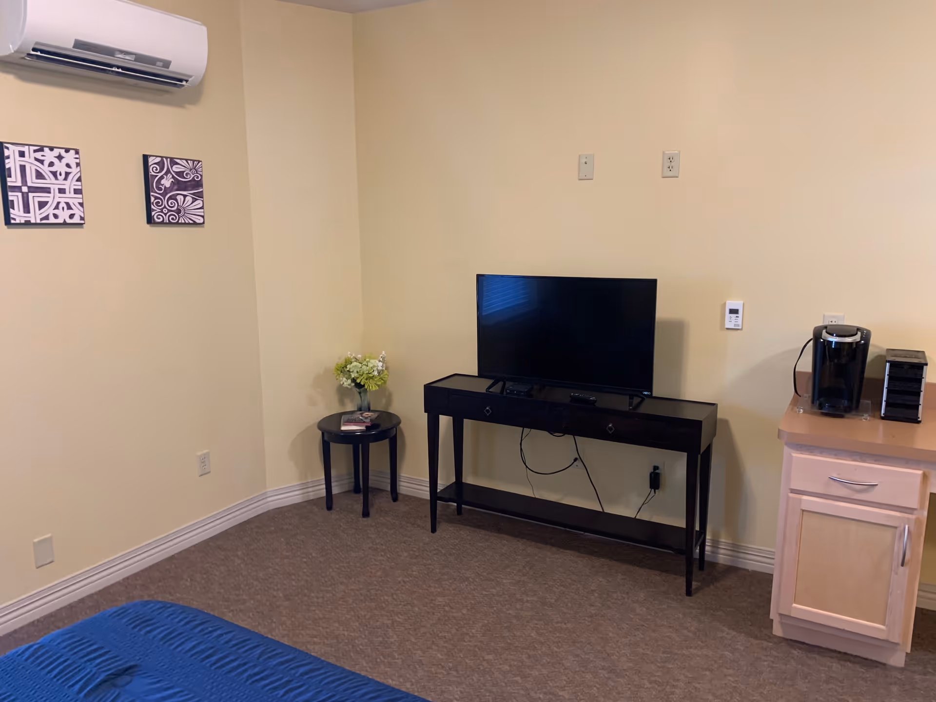 A corner of a room with beige walls and carpeted floor. There is a small black table with a vase of flowers and a magazine, a black console table with a flat-screen TV on it, and a light wood cabinet with a coffee maker and a small black organizer on top. Two decorative wall art pieces hang on the left wall, and an air conditioning unit is mounted above them.