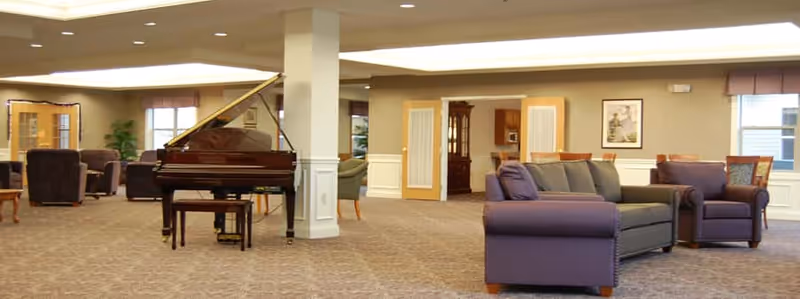 Spacious assisted living common area with a grand piano in the center, several upholstered chairs and sofas arranged around the room, beige walls with white wainscoting, and large windows letting in natural light.