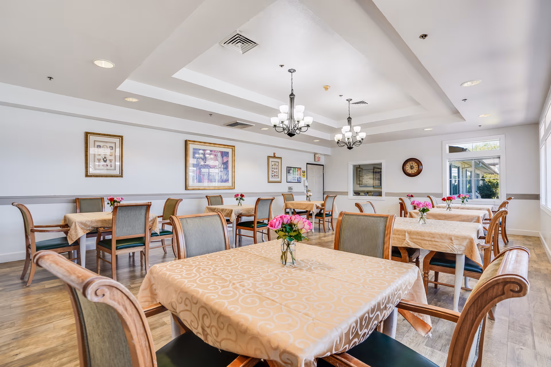 Bright dining room with multiple tables covered in beige tablecloths, wooden chairs, chandeliers, and small vases of flowers.