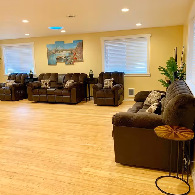 A spacious living room with light wooden flooring and yellow walls. The room features several dark brown cushioned sofas and armchairs with patterned pillows. There are two windows with white blinds, a small round wooden side table, two black side tables with decorative items, a large green plant, and a multi-panel wall art depicting a scenic view.