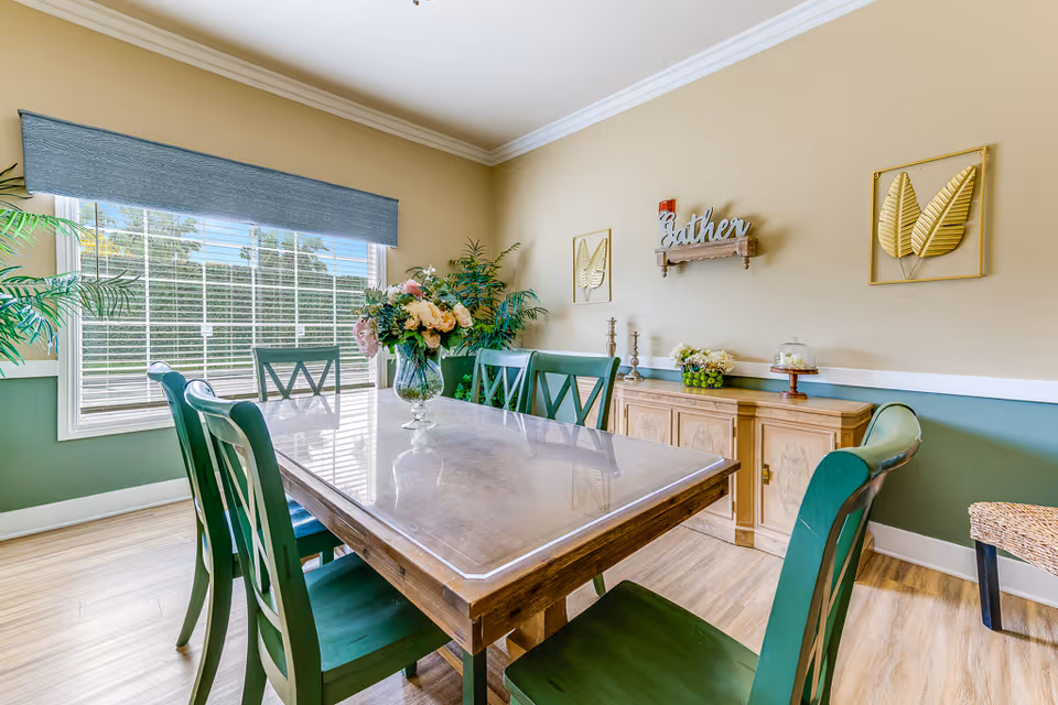 A bright dining room with a wooden table and six green chairs. A large window with white blinds and a blue valance lets in natural light. The walls are painted beige and green with white trim. On the wall, there are two gold leaf decorations and a wooden shelf with the word 'Gather' displayed. A wooden sideboard holds decorative items including flowers and candlesticks. There are also green plants near the window.