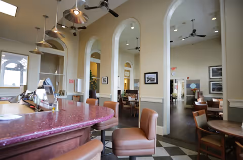 Interior view of a senior living facility dining area with a curved pink countertop bar and several brown cushioned bar stools. The room features high ceilings with ceiling fans and pendant lights, arched doorways leading to additional dining spaces with tables and chairs, and framed artwork on the walls.