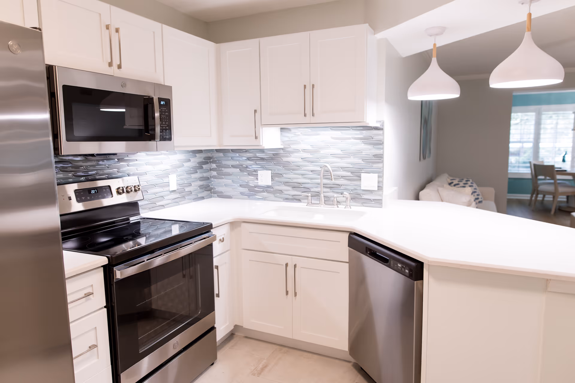 Bright modern kitchen with stainless steel appliances, white cabinets, a mosaic tile backsplash, and a curved white breakfast bar opening to a living area.