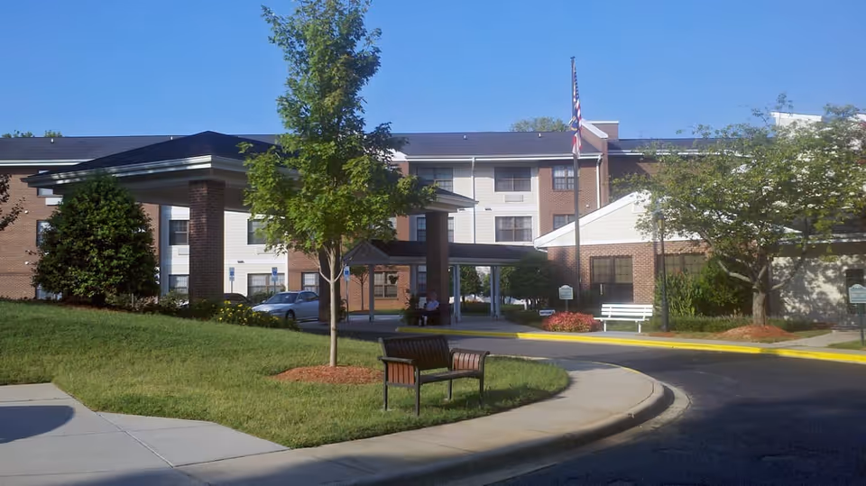 Front exterior of a senior living building with a covered entrance, benches, landscaping, and an American flag.