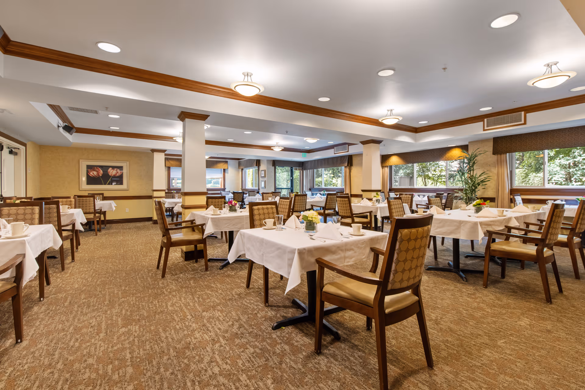 A spacious dining room with multiple tables covered in white tablecloths, each set with cups, napkins, and small flower arrangements. The room has large windows allowing natural light, beige walls, carpeted floor, and wooden chairs with cushioned seats and backs. Ceiling lights and wooden trim add warmth to the space.