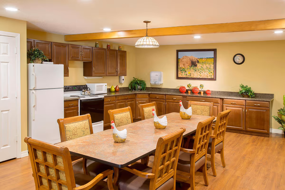 Communal dining area with a long table and chairs in front of a fully equipped kitchen with wooden cabinets and appliances.