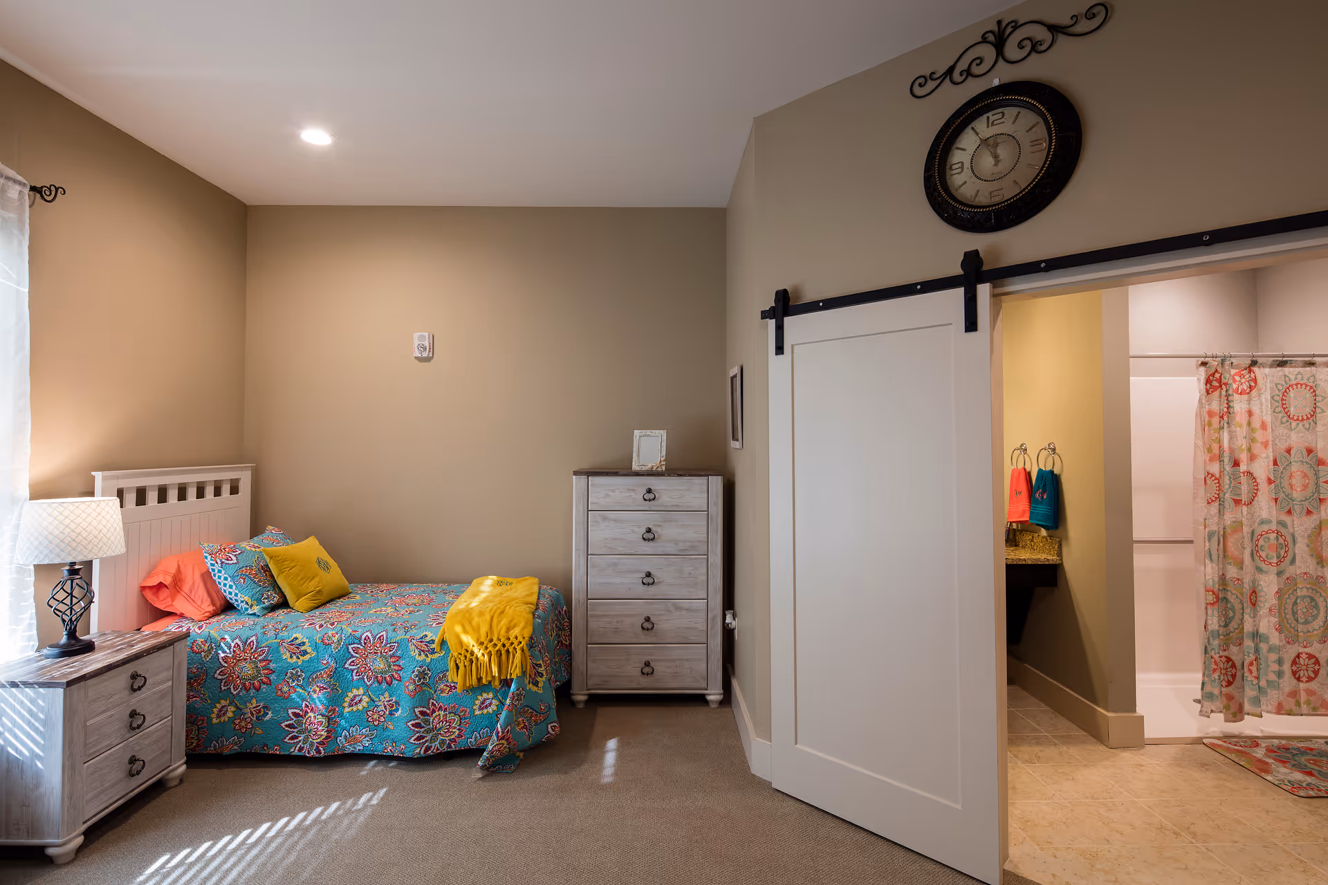 A cozy bedroom with a single bed covered in a colorful floral bedspread and multiple pillows. Next to the bed is a nightstand with a lamp. Across from the bed is a tall dresser. A sliding barn door leads to a bathroom with a shower curtain featuring a circular pattern and colorful towels hanging near the sink.