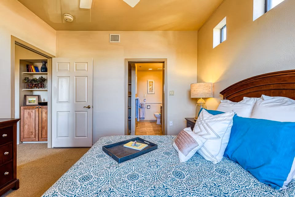 A furnished bedroom with a patterned blue bedspread, decorative pillows, a bedside lamp, and an open doorway leading to a bathroom.