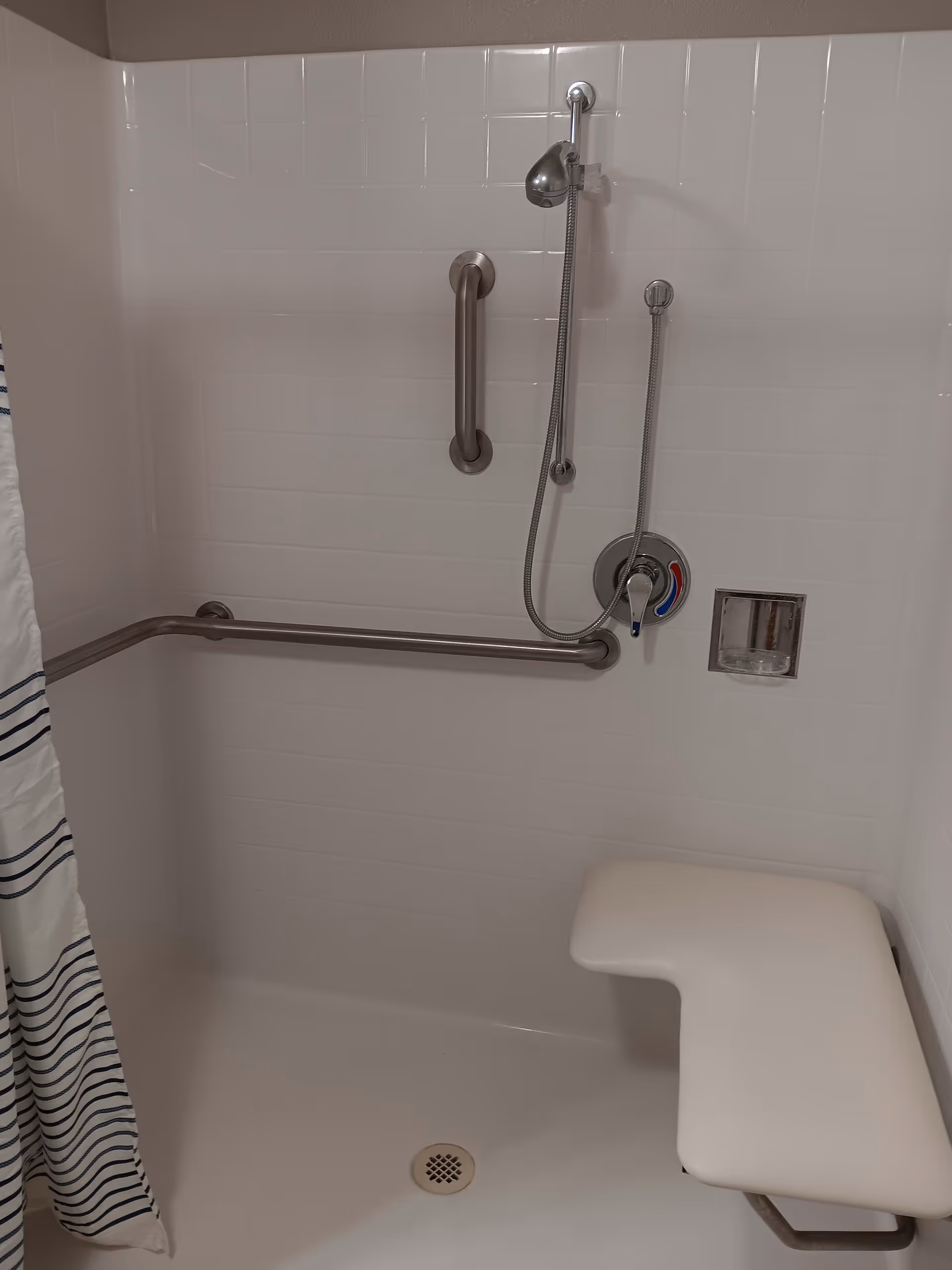 A white tiled shower area with a handheld showerhead, two stainless steel grab bars, a built-in soap dish, and a white shower bench. A white shower curtain with blue stripes is partially visible on the left side.