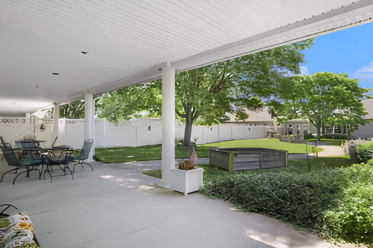 Covered outdoor patio area with several metal tables and chairs, overlooking a green lawn with trees and shrubs. The patio has white pillars and a white ceiling, and the surrounding area includes a white fence and residential buildings in the background under a clear blue sky.