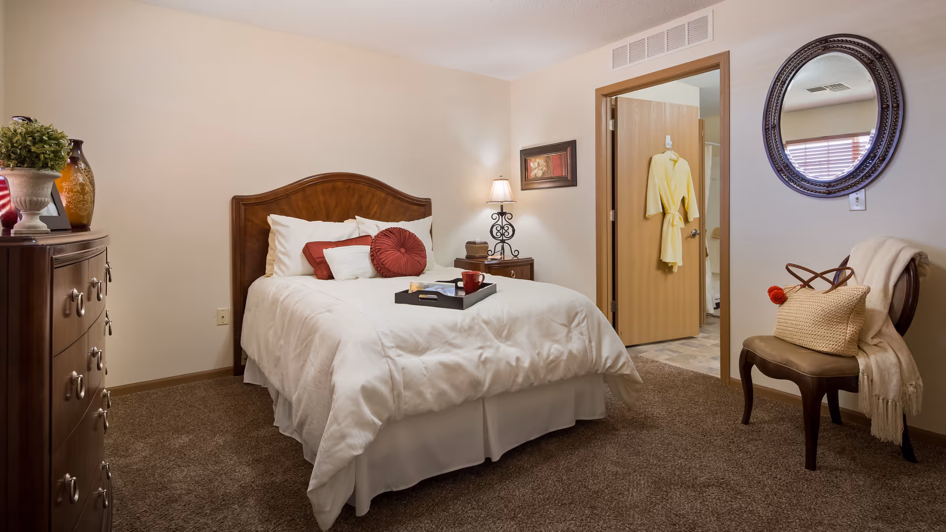 A cozy bedroom with a wooden bed frame and white bedding accented by red pillows. A wooden dresser with decorative items is on the left, and a nightstand with a lamp and a framed picture is beside the bed. An open door reveals a glimpse of a bathroom with a yellow robe hanging on the door. A chair with a beige throw and a woven bag sits beneath a round mirror on the right wall.