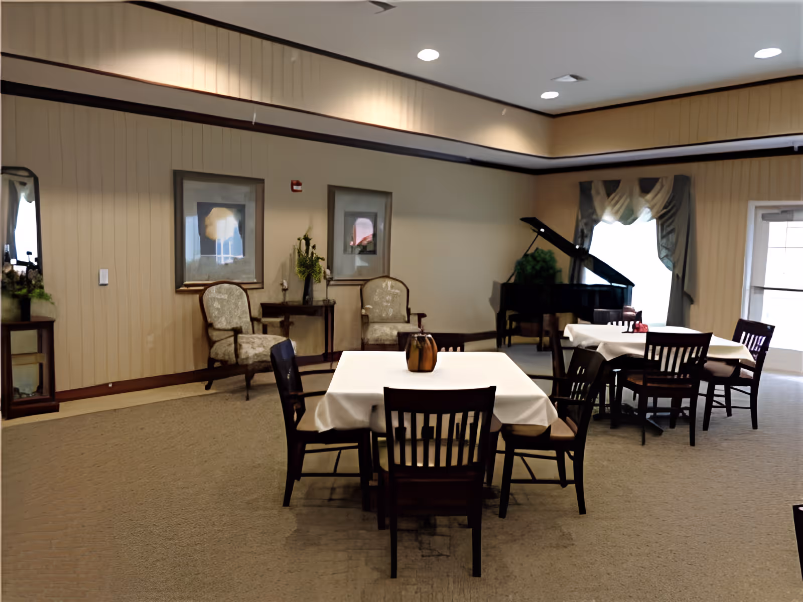 Interior of a senior living community common area with two square tables covered with white tablecloths, each surrounded by wooden chairs. In the background, there are two upholstered armchairs with a small table between them, two framed pictures on the wall, a grand piano near a window with draped curtains, and a door leading outside.