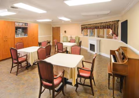Dining room with tables covered in white cloths, red chairs, and a piano along the wall.