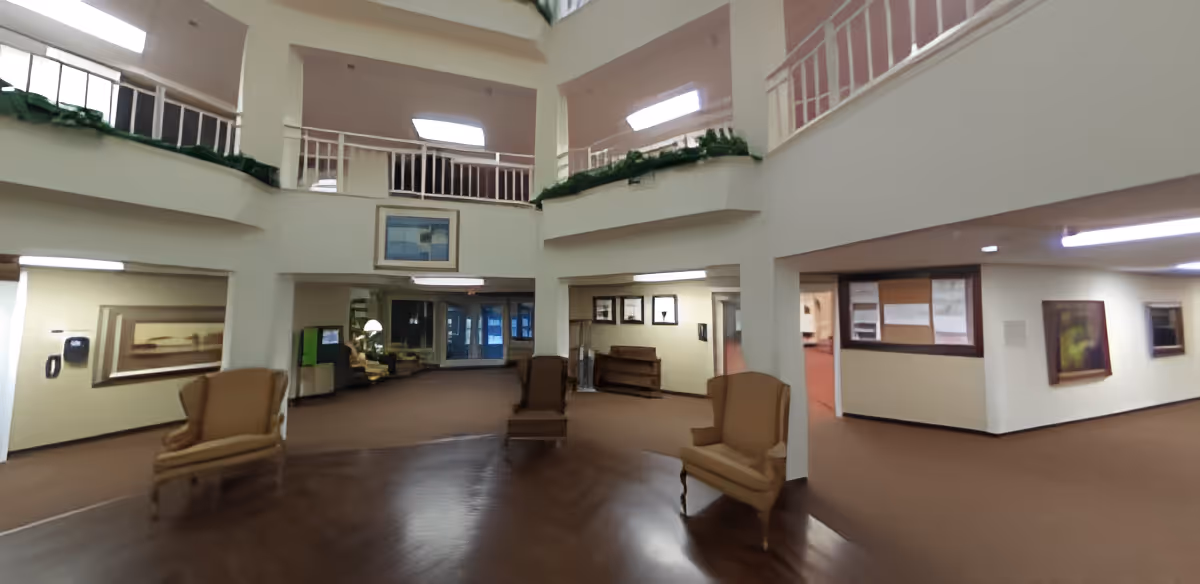 Interior view of a senior living facility lobby or common area with high ceilings and an upper balcony. The space features several armchairs arranged around the room, framed artwork on the walls, and a mix of carpet and wood flooring. There are windows and doors leading to other rooms and areas within the facility.