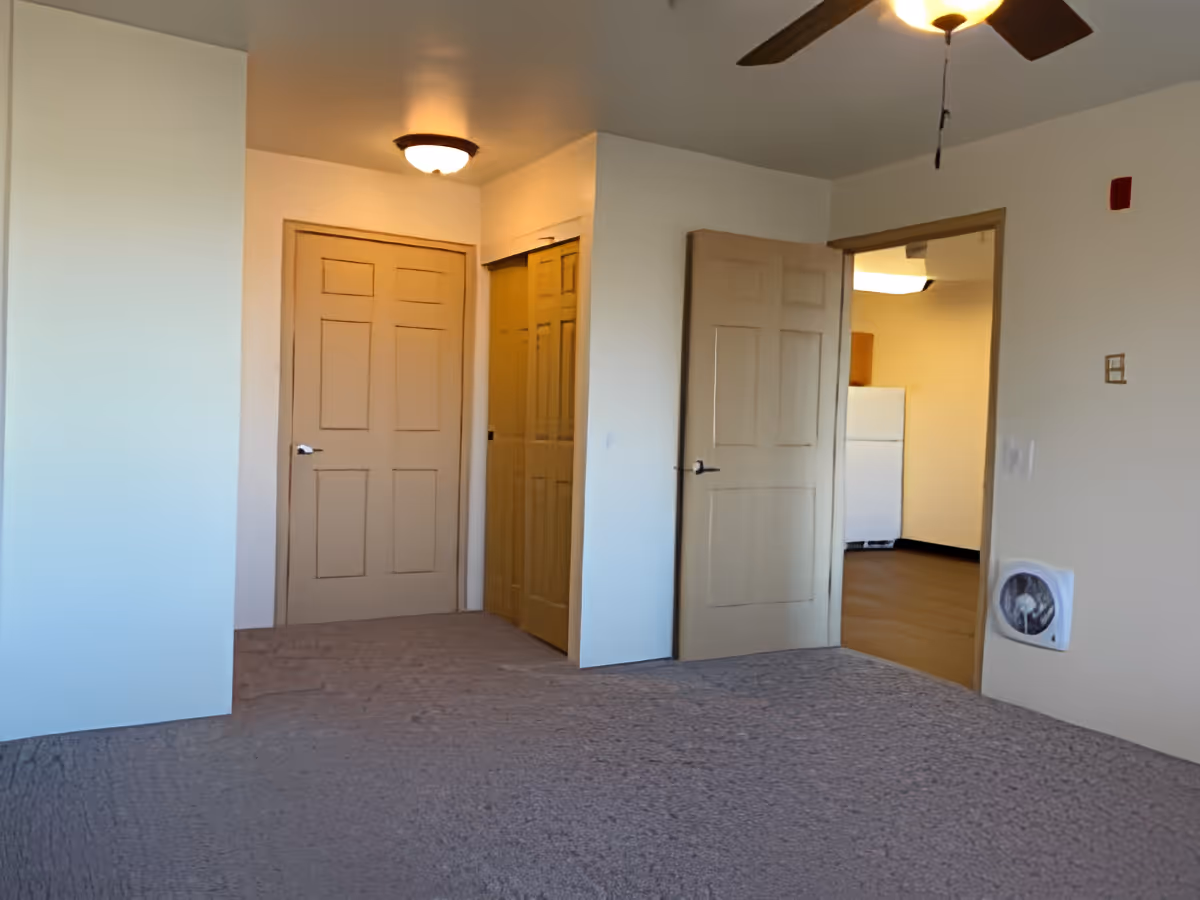Interior view of a room with beige walls and carpeted floor, featuring three doors including a closet with sliding doors and an open door leading to a kitchen area with a white refrigerator. A ceiling fan with a light is partially visible on the ceiling.