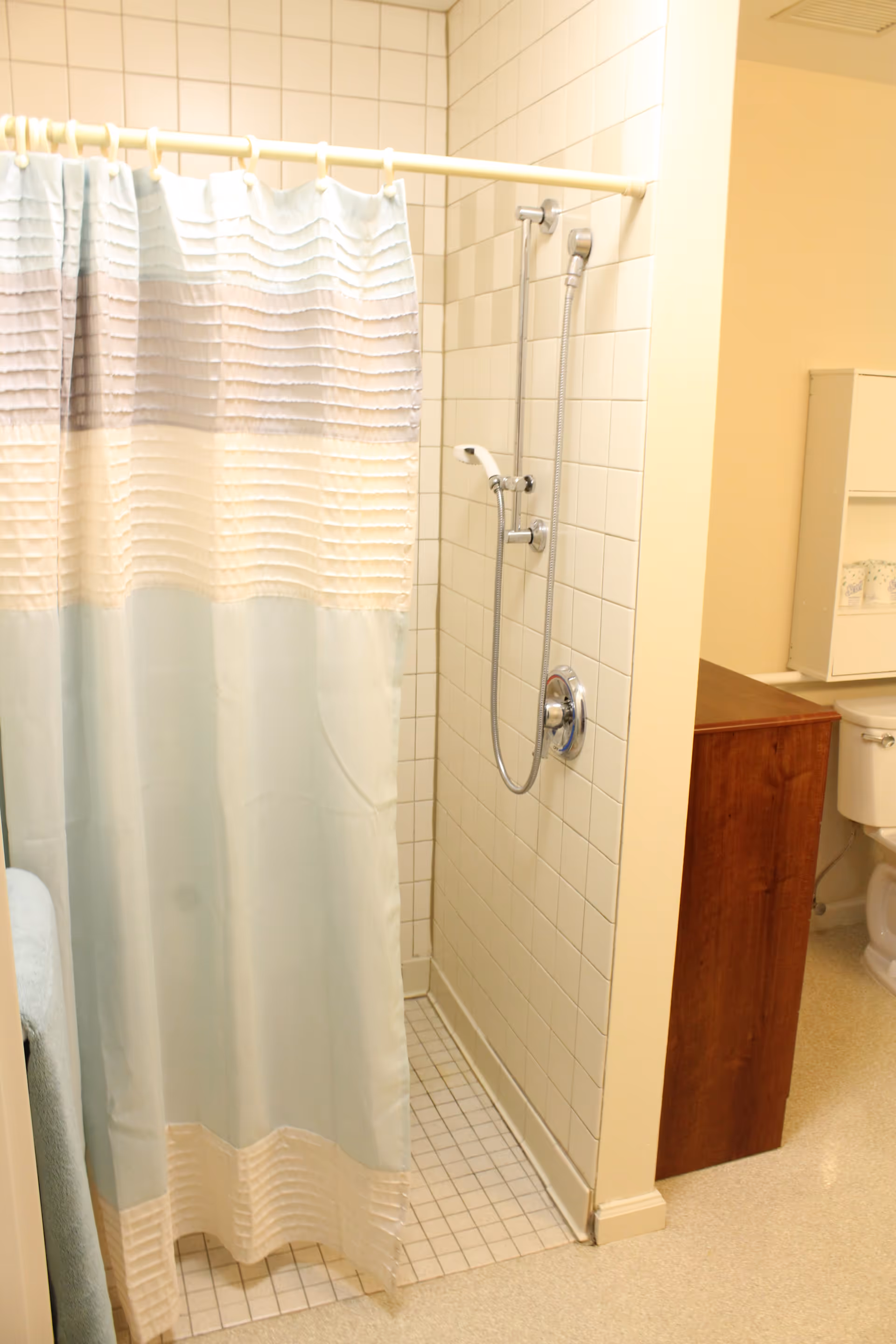 A bathroom shower area with white tiled walls and floor, featuring a handheld showerhead mounted on the wall. A light blue and white striped shower curtain is partially closed. To the right, a wooden cabinet and a white toilet are visible.