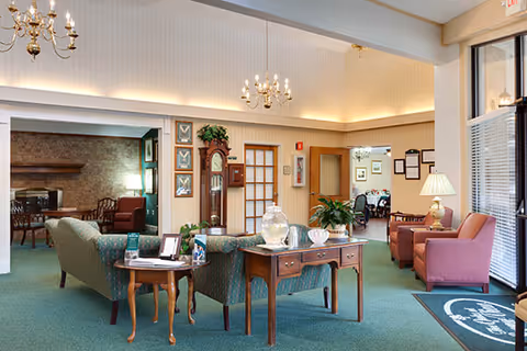 A spacious and well-lit assisted living facility common area with green carpet and cream-colored walls. The room features comfortable seating including a green patterned sofa, pink armchairs, and wooden tables with decorative items such as plants, a glass jar, and framed photos. Two chandeliers hang from the ceiling, and there is a grandfather clock against the wall. Large windows with blinds allow natural light to enter, and adjoining rooms with additional seating are visible in the background.