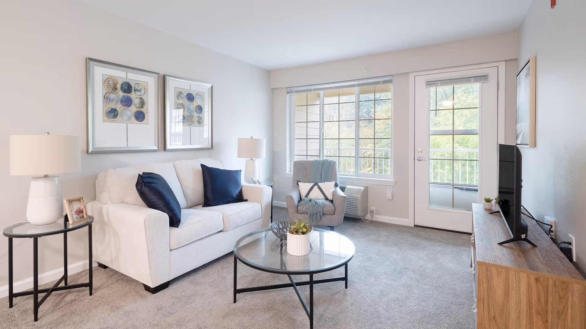 A bright and cozy living room with a white sofa adorned with two dark blue pillows, a gray armchair with a blue throw blanket, a round glass coffee table with a small plant, two framed abstract artworks on the wall, a wooden TV stand with a flat-screen TV, and large windows and a glass door letting in natural light.