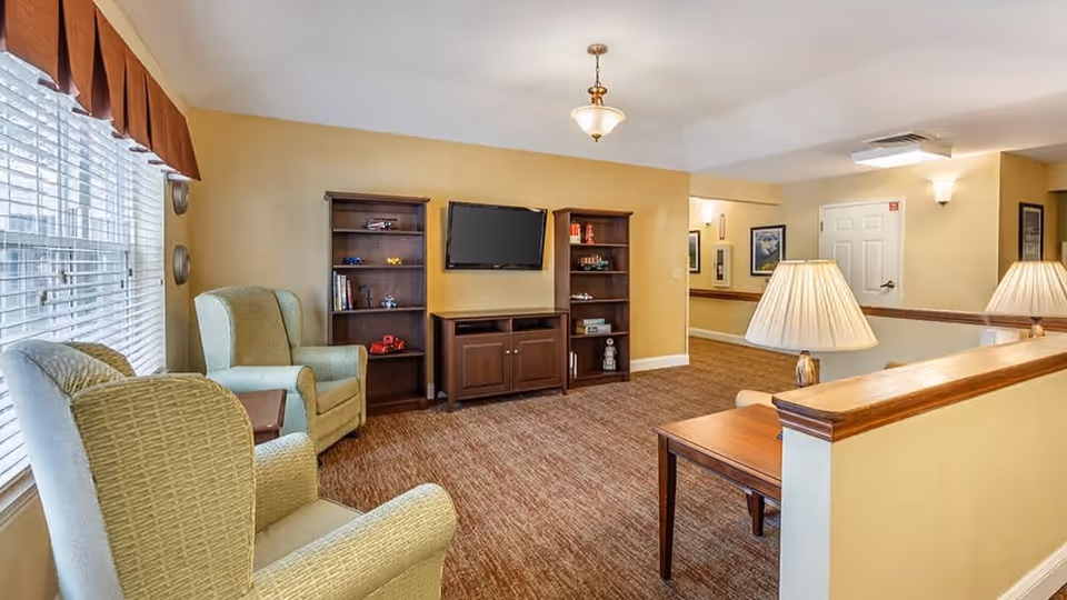 A cozy living room area in a senior living facility with two cushioned armchairs near a large window with blinds and a valance. There are two wooden bookshelves and a TV mounted on the wall between them. A wooden side table with a lamp is in the foreground, and the walls are painted a warm beige color with carpeted flooring.