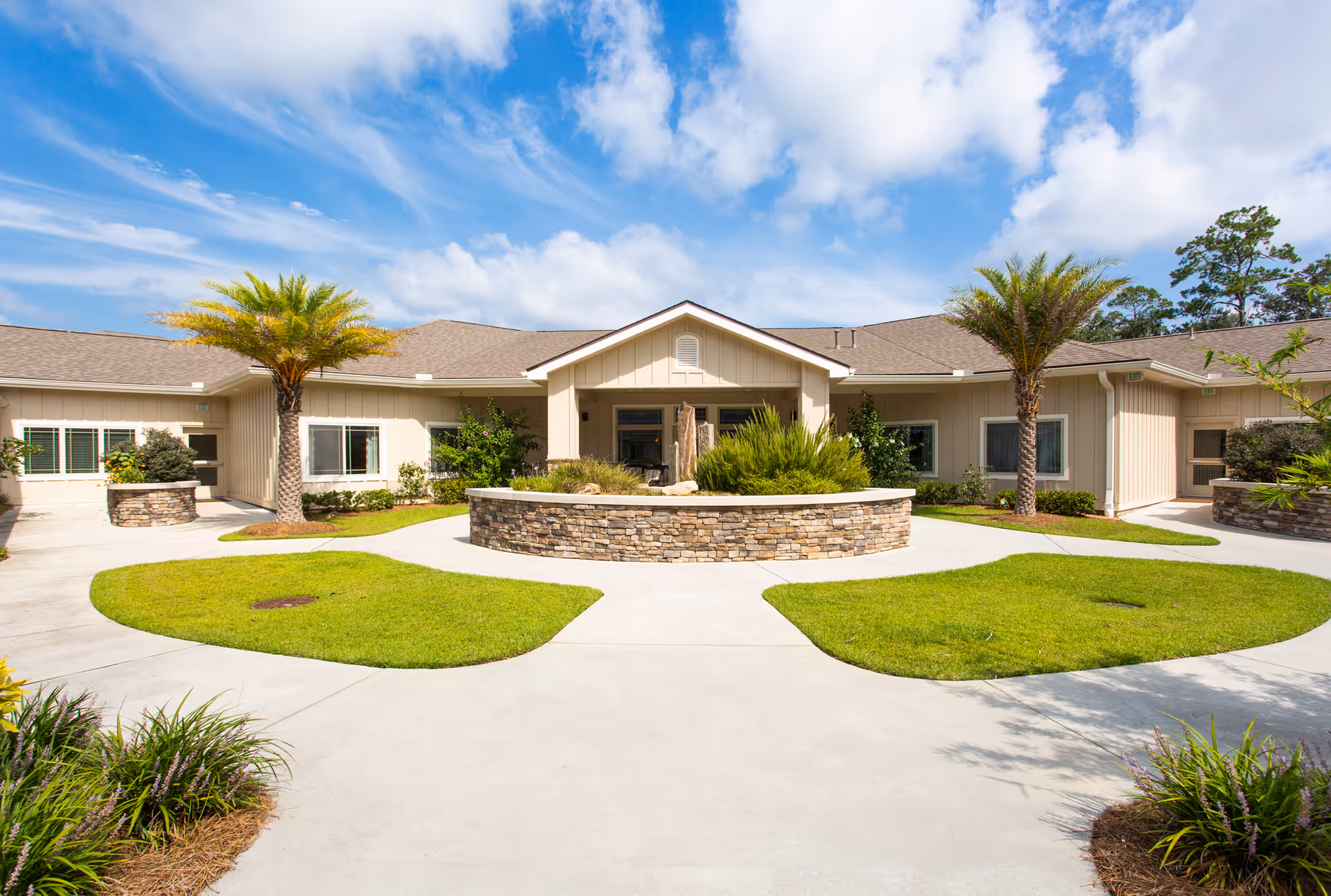 Exterior view of a single-story assisted living facility building with beige siding, a central entrance, and a landscaped courtyard featuring palm trees, green grass, and a stone planter under a partly cloudy blue sky.