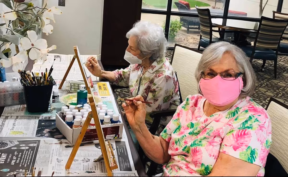 Two elderly women wearing face masks are seated at a table indoors, engaged in painting on small easels. The table is covered with newspapers and various paint bottles and brushes. One woman is wearing a pink floral shirt and a pink mask, while the other has white hair and is wearing a butterfly-patterned shirt and a white mask. Behind them, large windows show an outdoor patio area with chairs and tables.