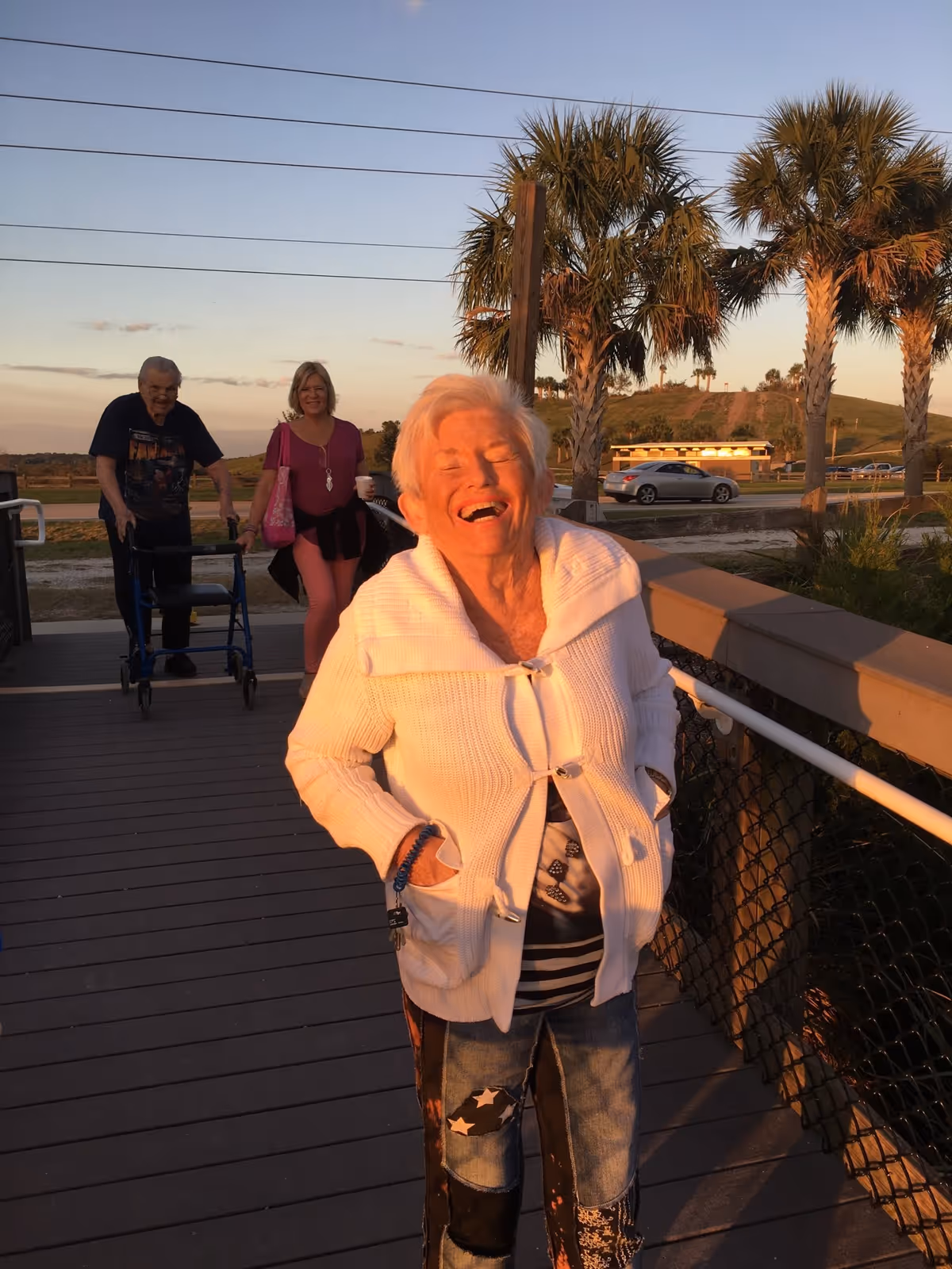 An elderly woman wearing a white sweater and patterned jeans is smiling and standing on a wooden boardwalk with her hands in her pockets. Behind her, a man using a walker and a woman carrying a pink bag are walking. There are palm trees, a road with cars, and a grassy hill in the background during sunset.